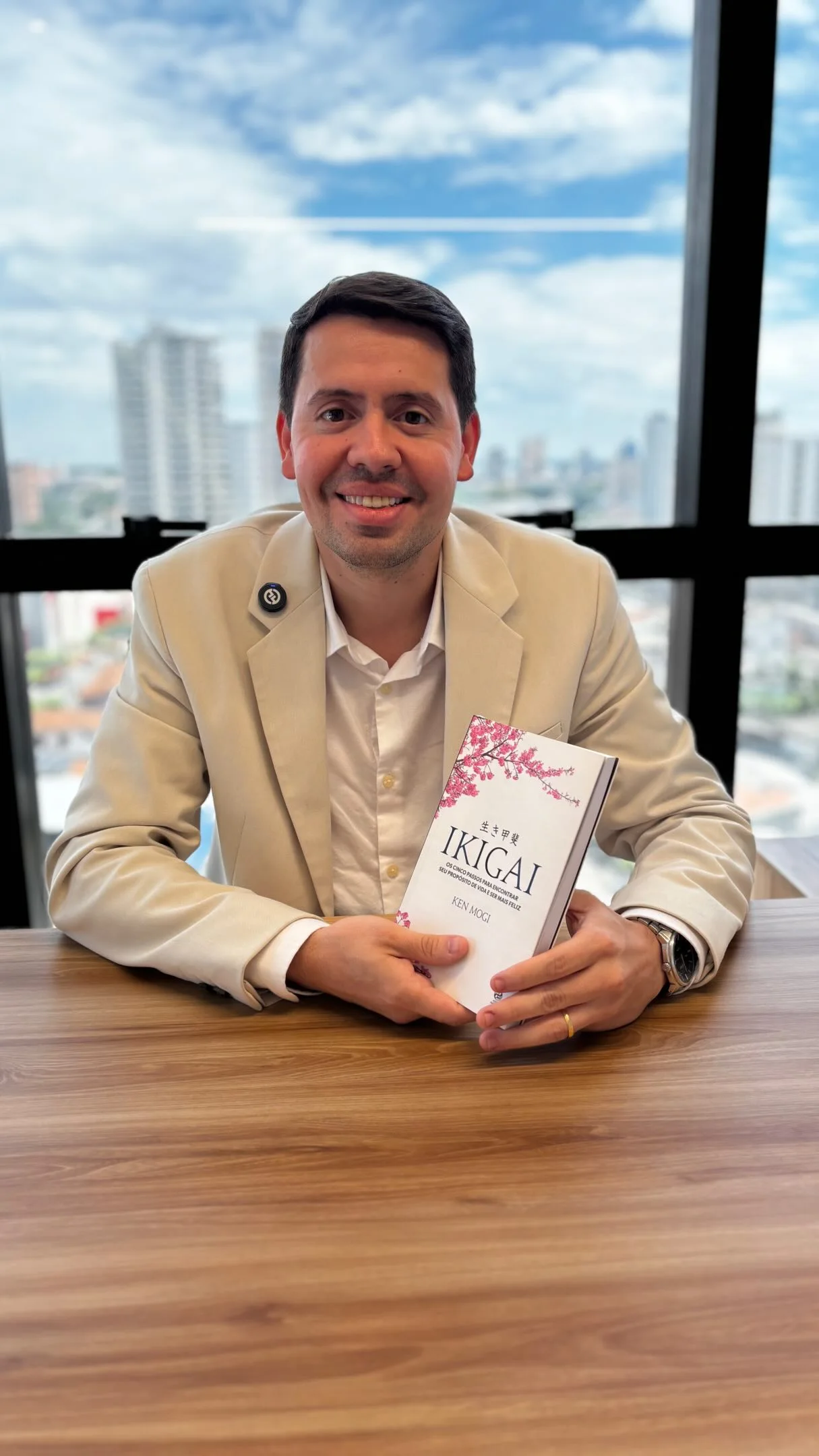 Dr. Caio Blanch sorrindo à mesa de madeira, de blazer bege, segurando o livro Ikigai, com a janela e os prédios de Belém ao fundo