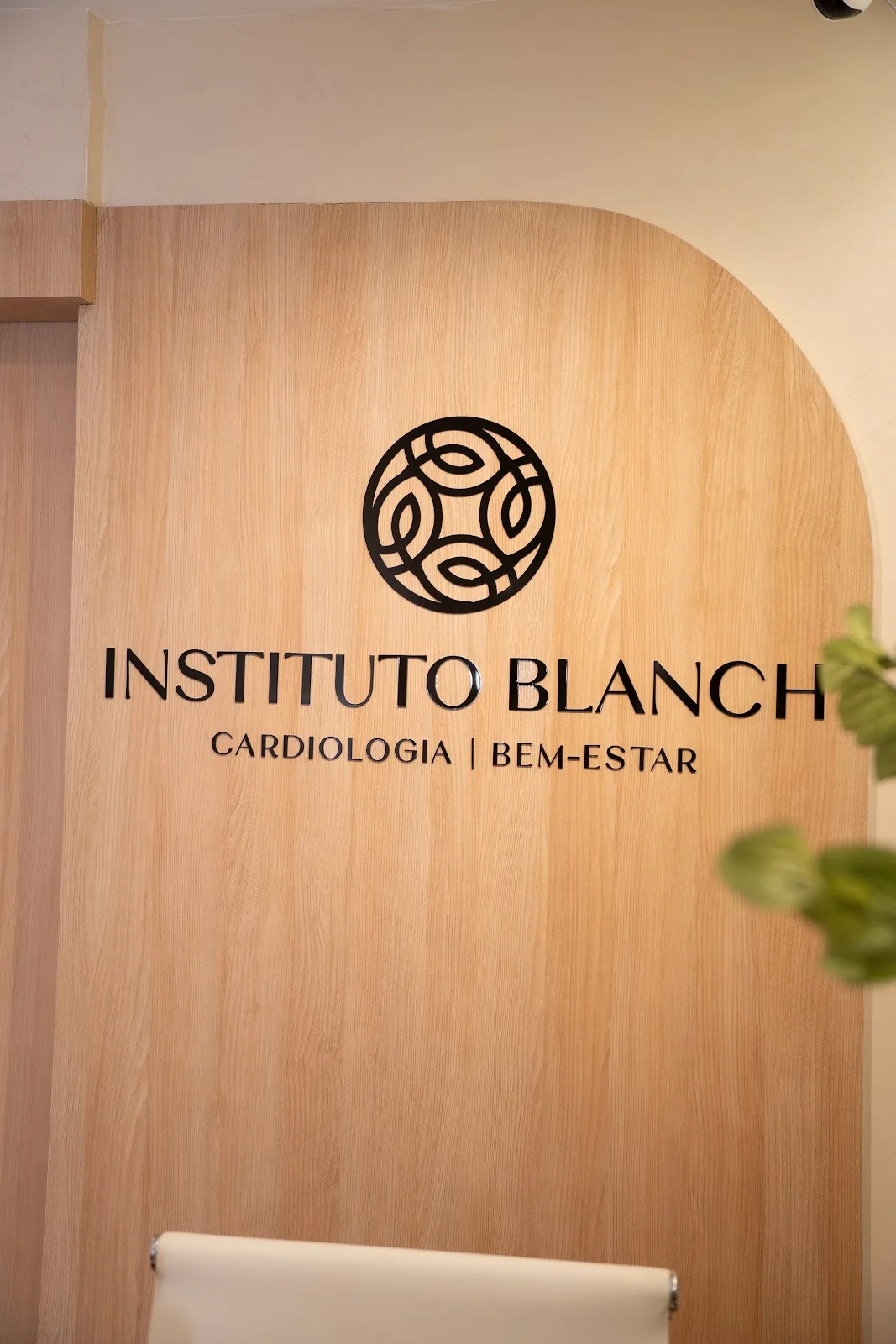 Recepção do Instituto Blanch: painel de madeira clara em arco com o letreiro em metal preto Instituto Blanch, Cardiologia e Bem-Estar, e uma poltrona branca à frente