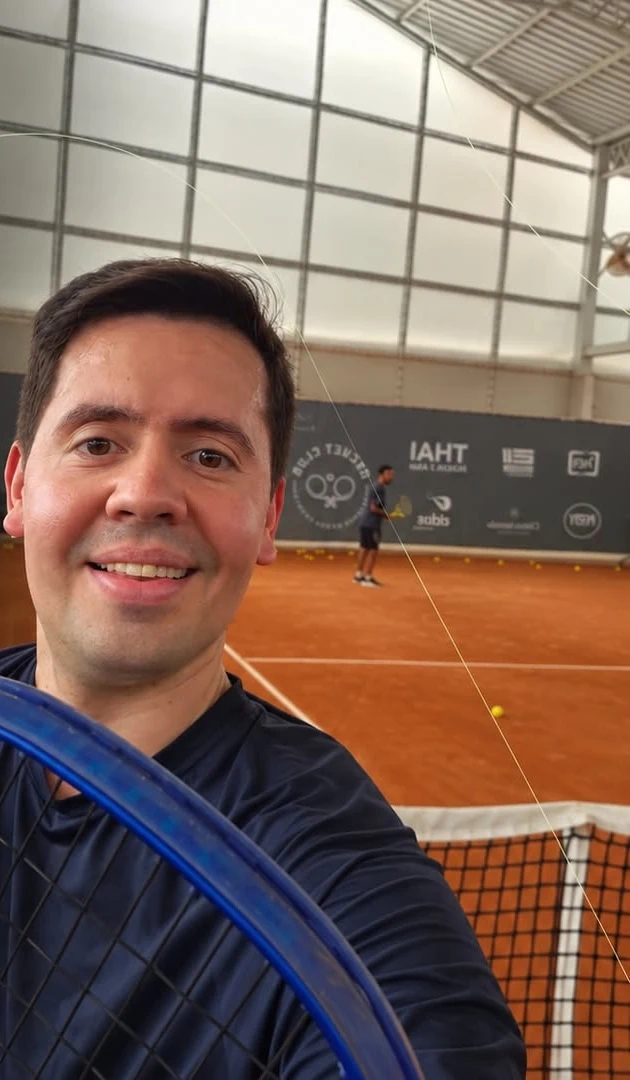 Dr. Caio Blanch sorrindo numa selfie em quadra de tênis de saibro coberta, com a raquete em primeiro plano